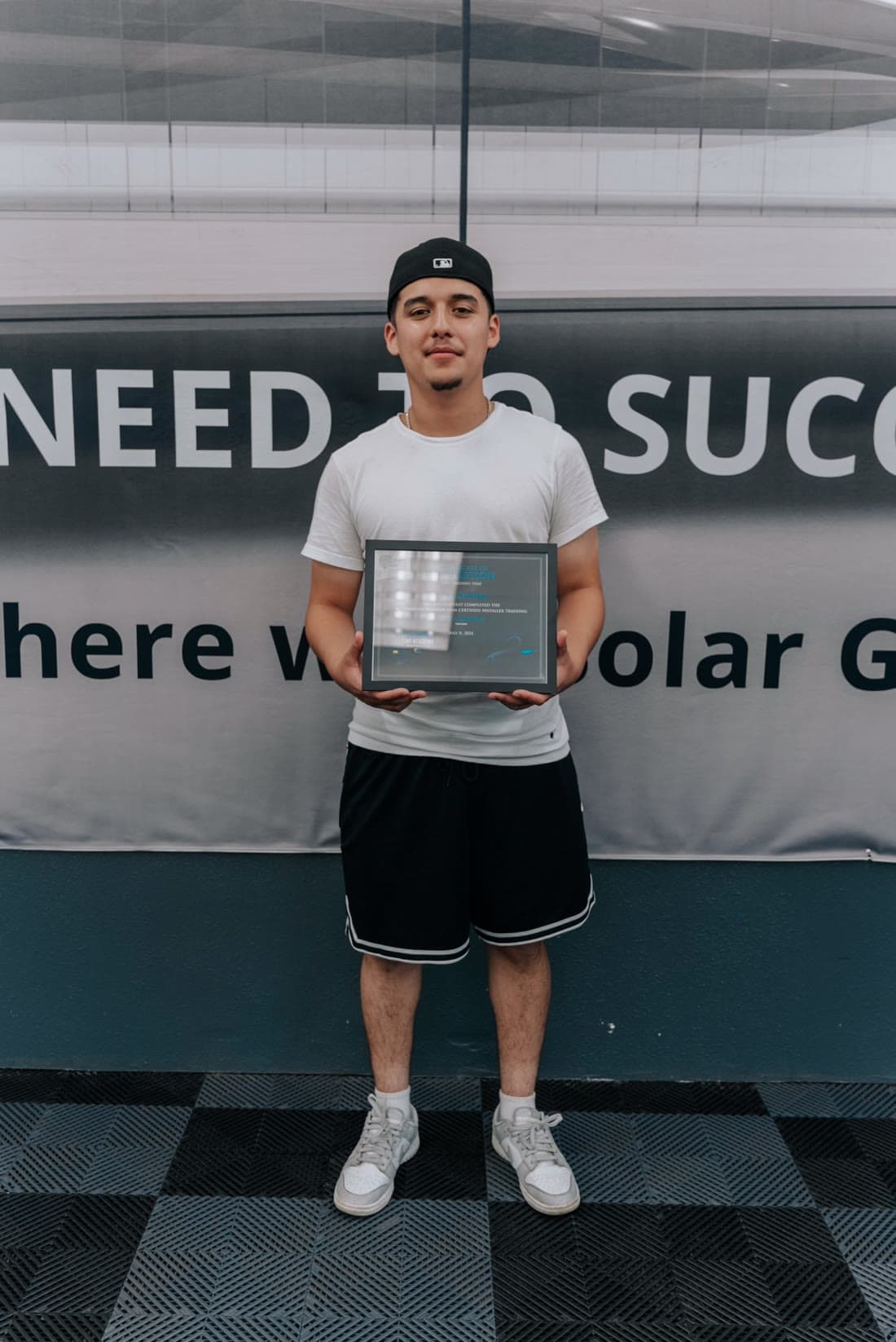 A graduate in a white T-shirt and cap holding his framed California Tint Academy certificate of completion in front of the Solar Gard wall
