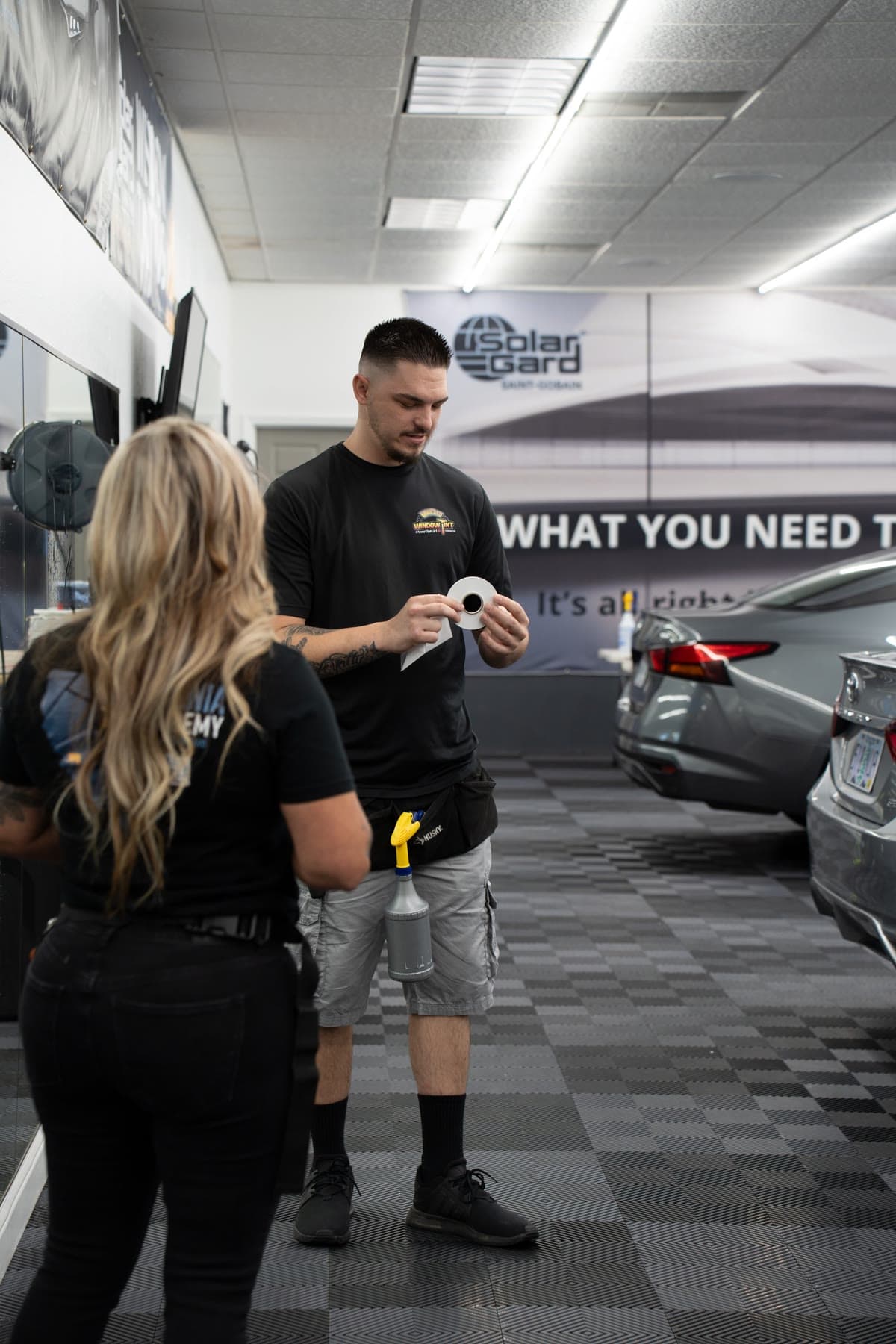 A student in a tool belt unrolling a fresh roll of window film while Roberta Rodriguez watches, in the Modesto training shop with the Solar Gard wall and training cars behind them
