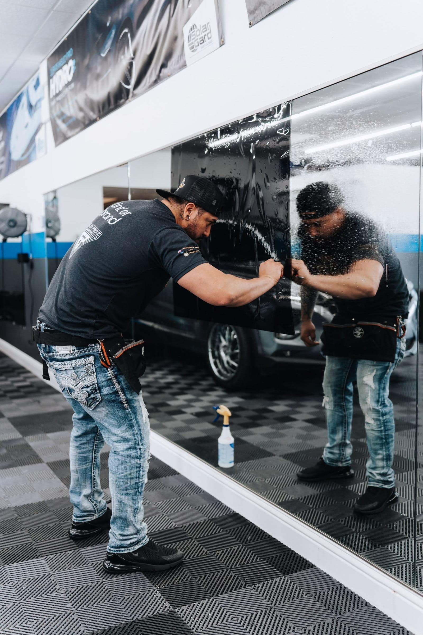 A California Tint Academy student squeegeeing dark window film onto a practice glass pane in the Modesto training shop, the shop reflected in the glass