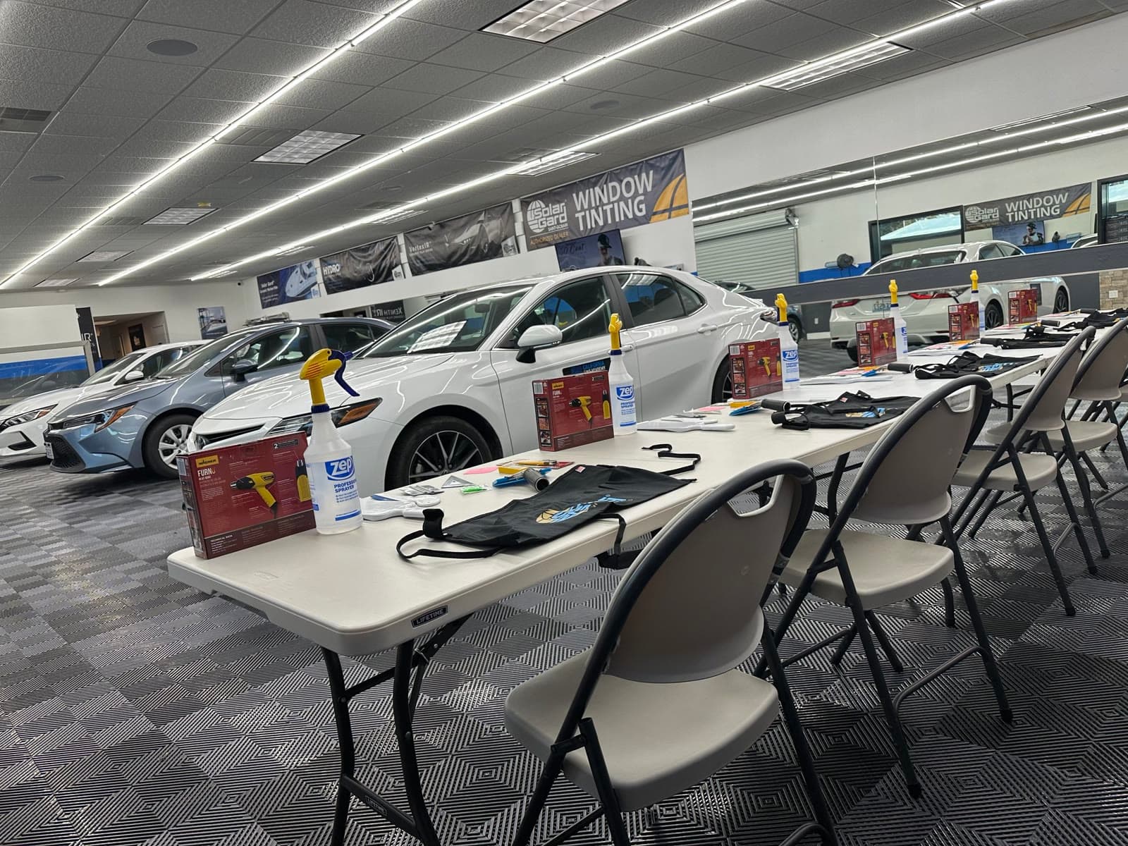 Rows of tool kits — heat guns, sprayers, aprons and hand tools — laid out at each seat on long tables in front of the training vehicles
